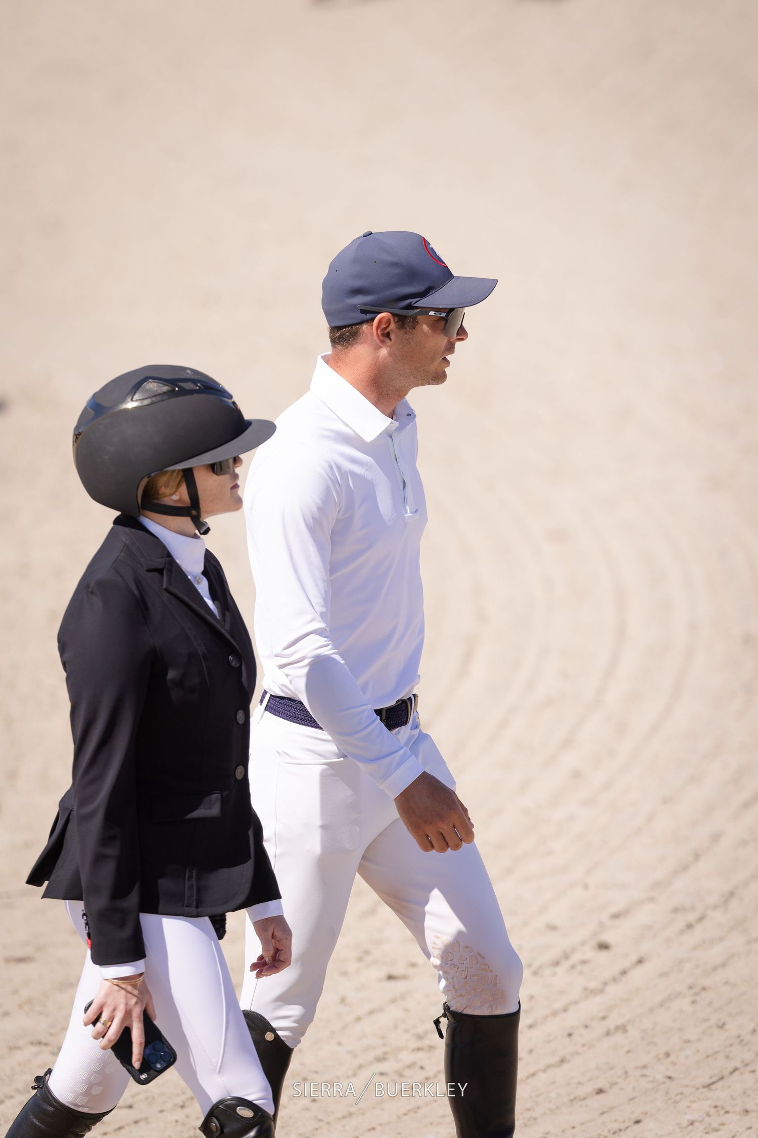Two equestrians in competition attire walking on sandy ground at a horse show.