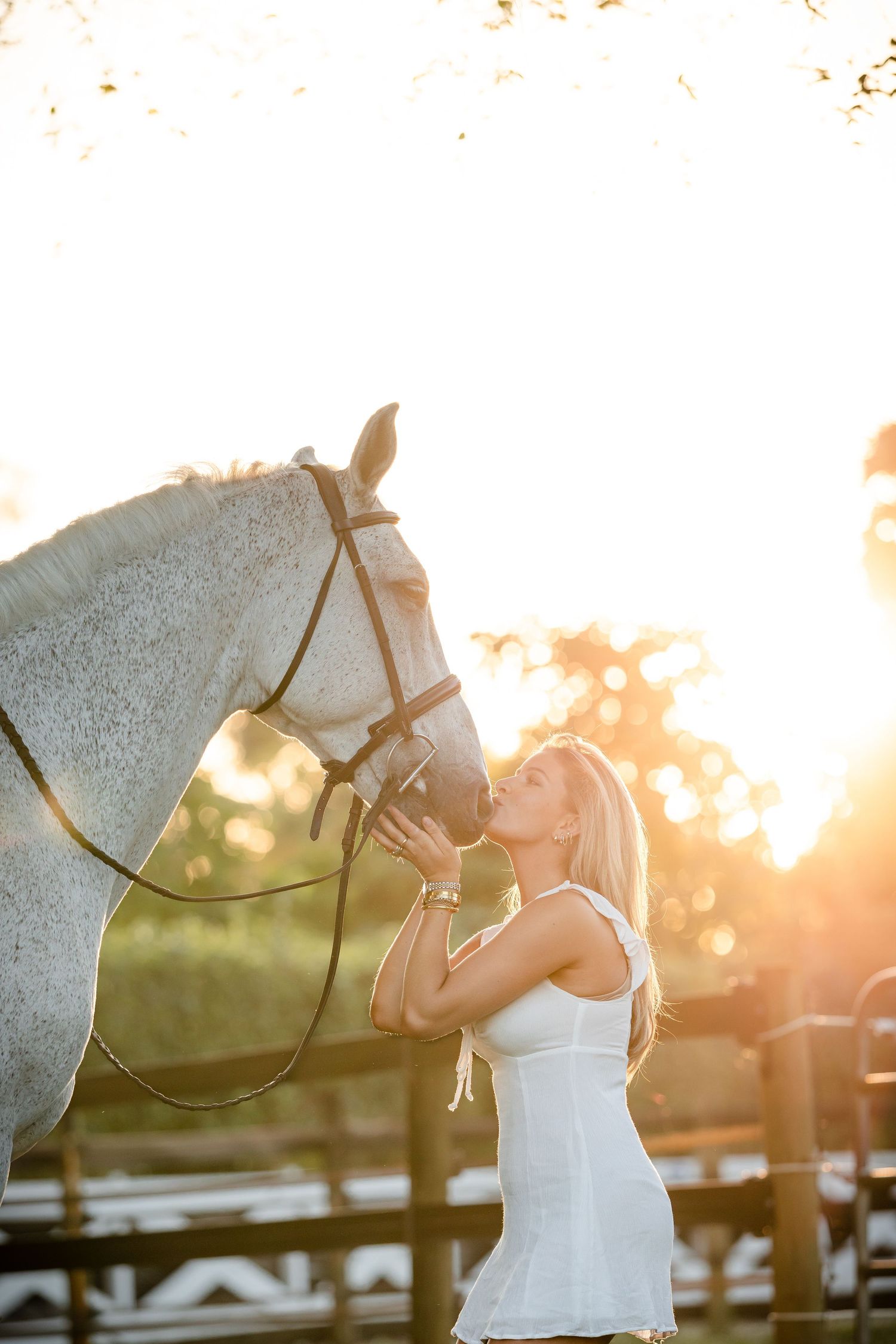A bride in a white dress lovingly embraces a horse in a rustic barn setting during golden hour sunset.