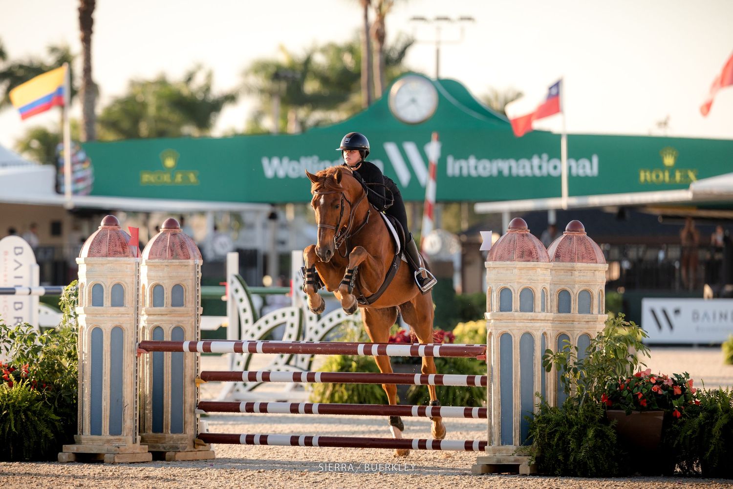 A rider and horse leap over a decorative jump barrier at an international equestrian competition during sunset.
