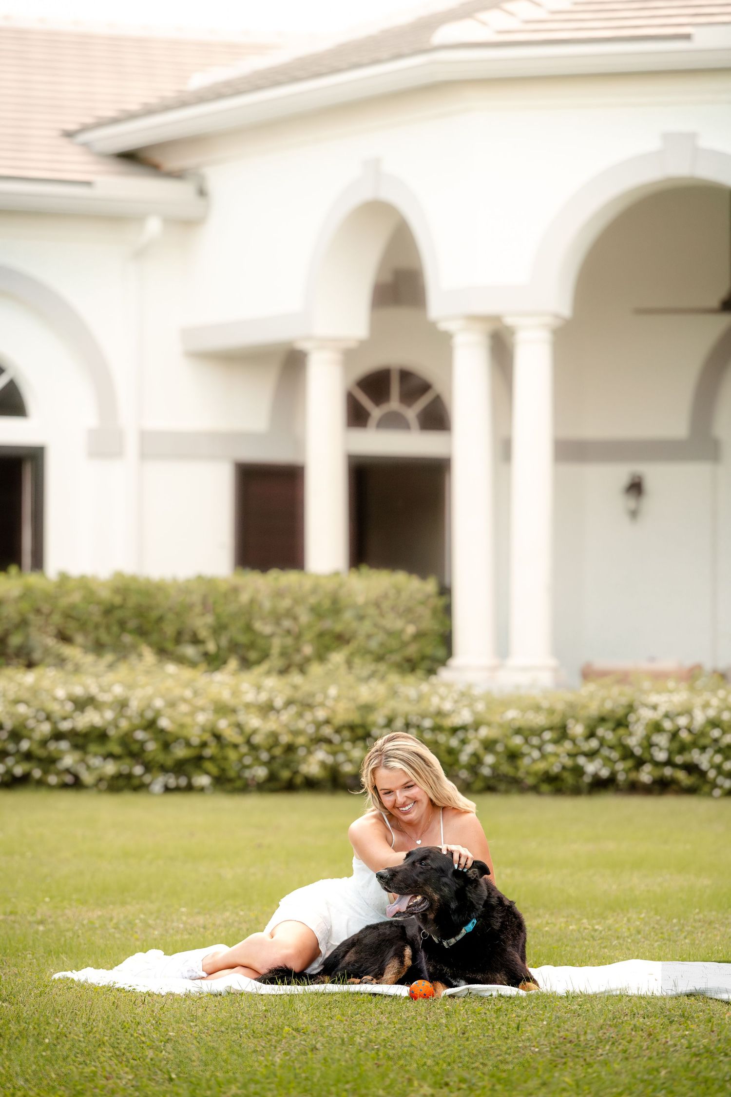 Woman relaxing on blanket with black dog on sunny lawn with white columned building in background.