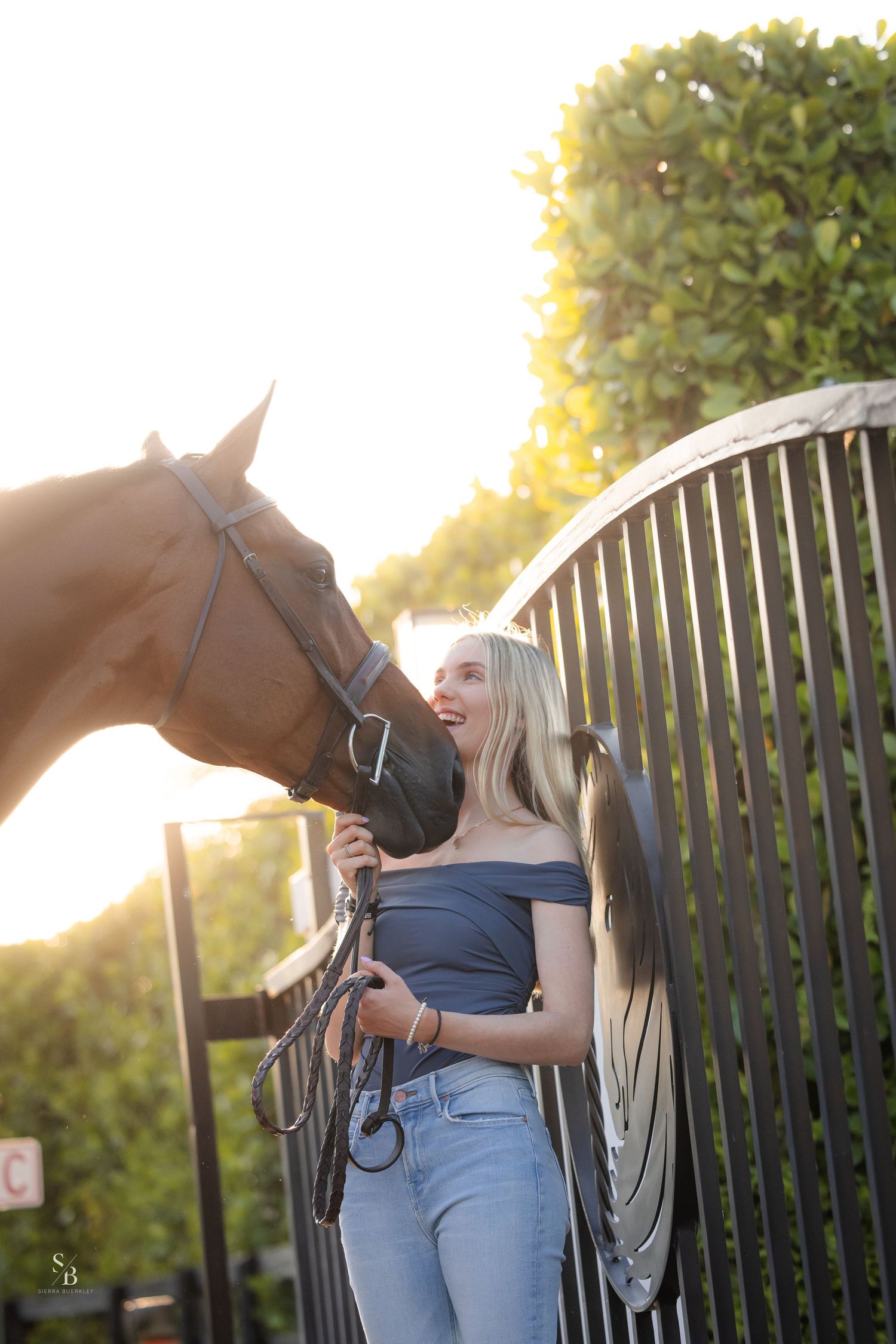A person stands with a brown horse against a black fence at sunset while wearing jeans and a tank top.