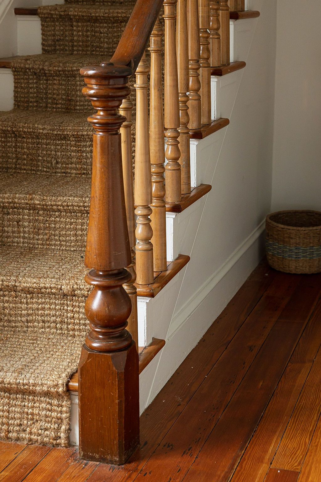 Close-up view of wooden staircase with beige sisal carpet runner and traditional turned balusters and newel post.