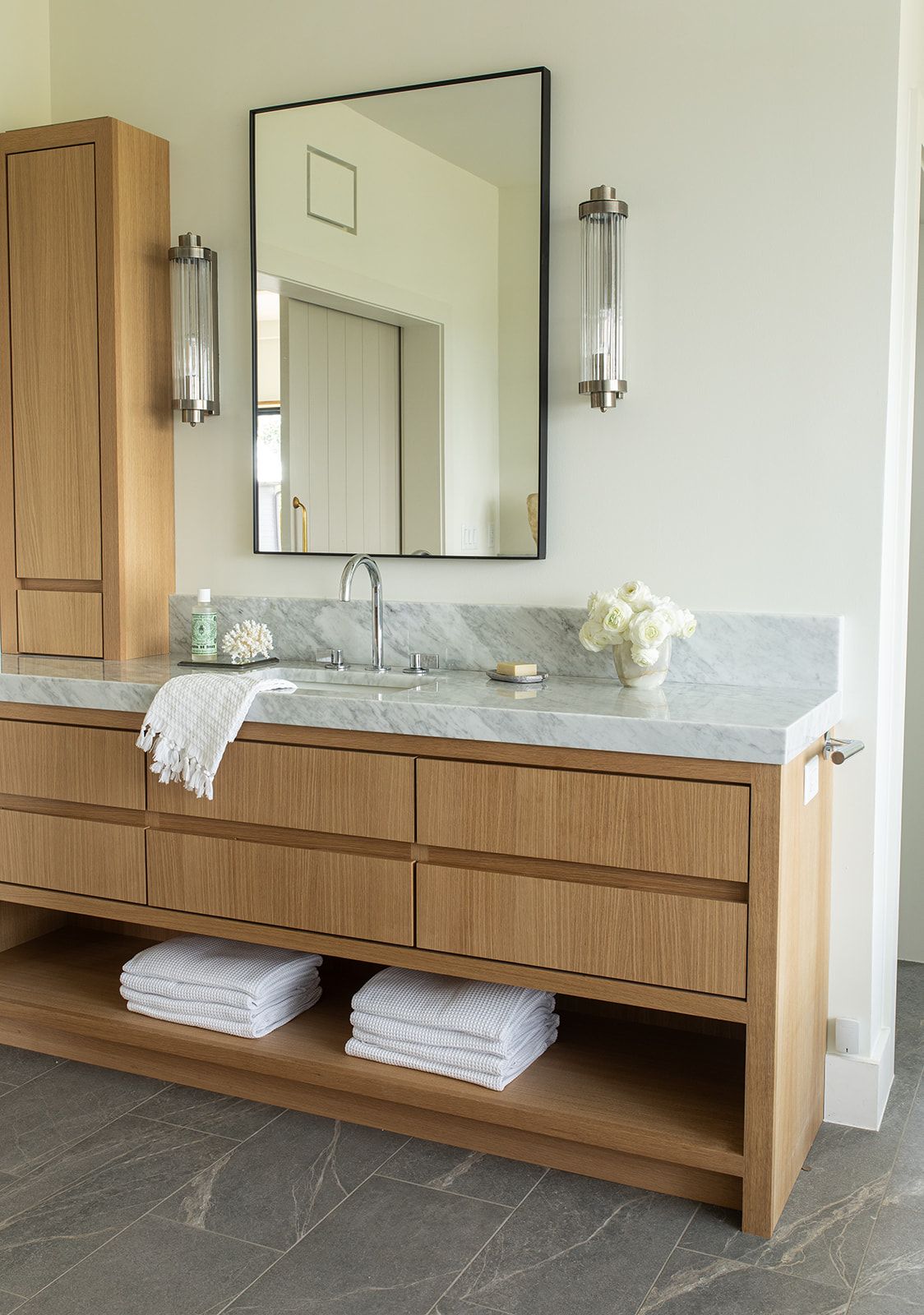 Natural oak bathroom vanity with marble countertop, black framed mirror, and brushed nickel wall sconces.