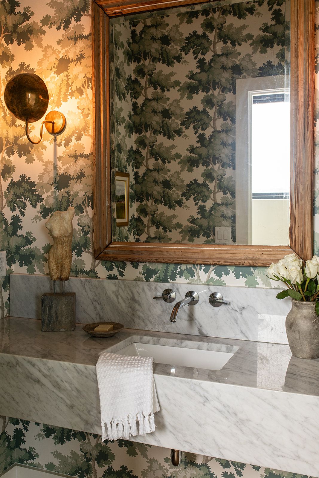 Elegant powder room with floral wallpaper, marble sink, gold-framed mirror, and brass wall sconce.