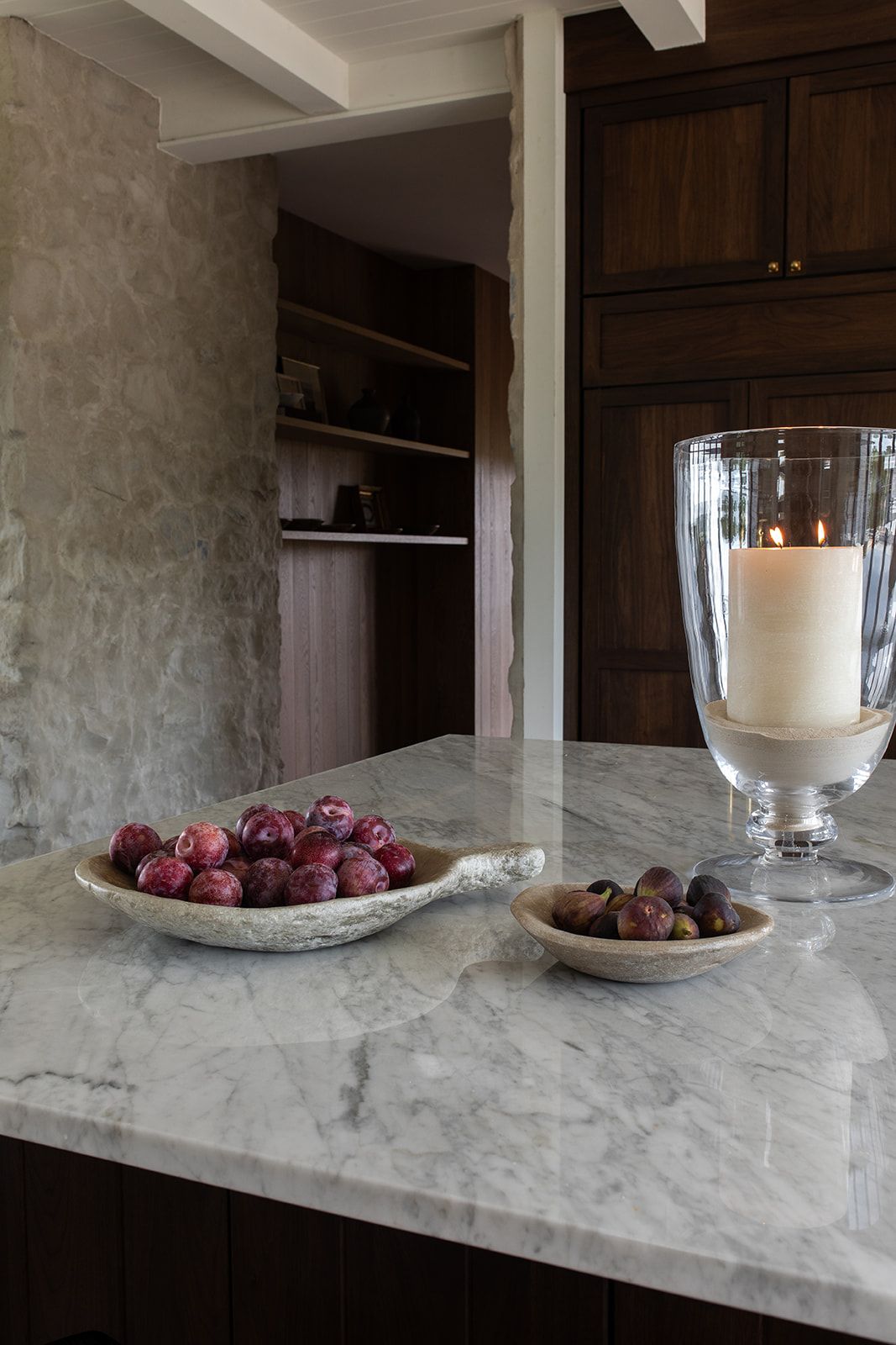 Marble kitchen island with fruit bowls and lit candle in glass hurricane, dark wood cabinetry and stone wall in background.
