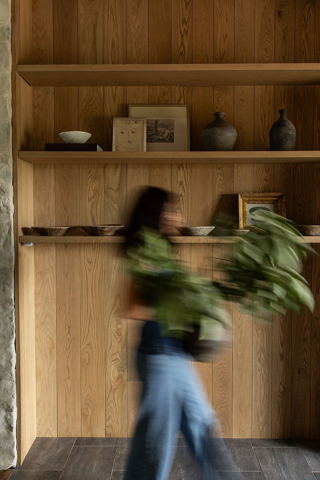 A blurred woman carries tropical leaves past wooden built-in shelves displaying vases, bowls, and framed art.