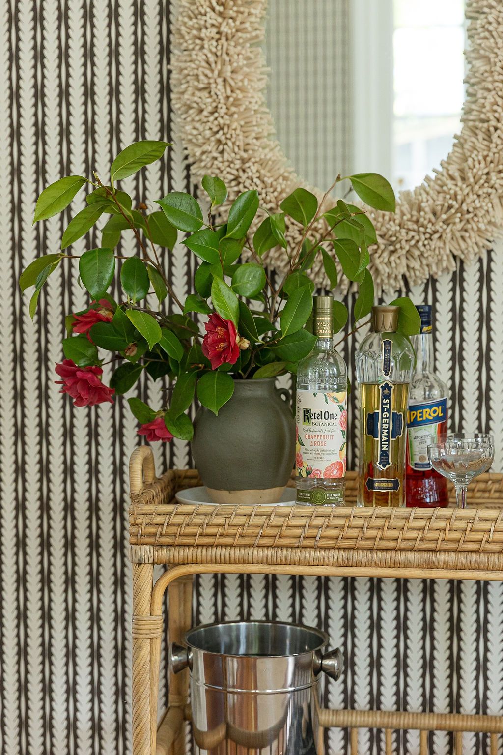 Modern home bar setup with rattan shelving, potted red flowers, and assorted liquor bottles displayed against striped wallpaper.