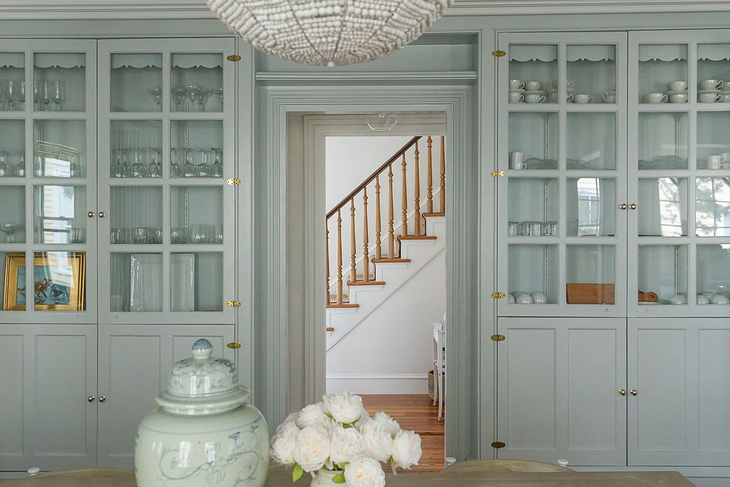 Elegant sage green built-in cabinets with glass doors frame a doorway leading to a white staircase beneath a crystal chandelier.