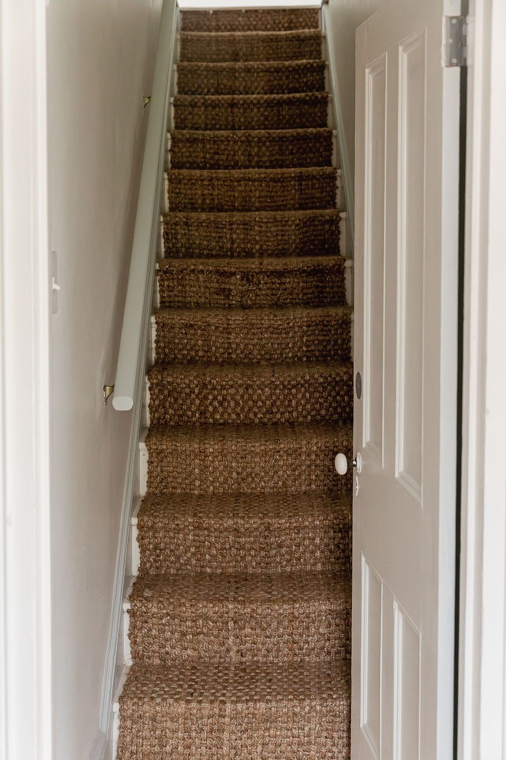 A narrow staircase with beige carpeting leads upward between white walls with a handrail mounted on one side.