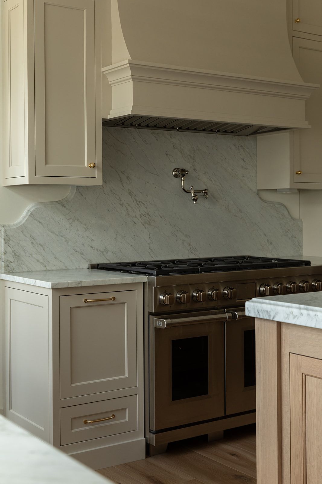 Elegant kitchen with taupe cabinets, marble backsplash, brass hardware, and a professional dark stainless range with pot filler.