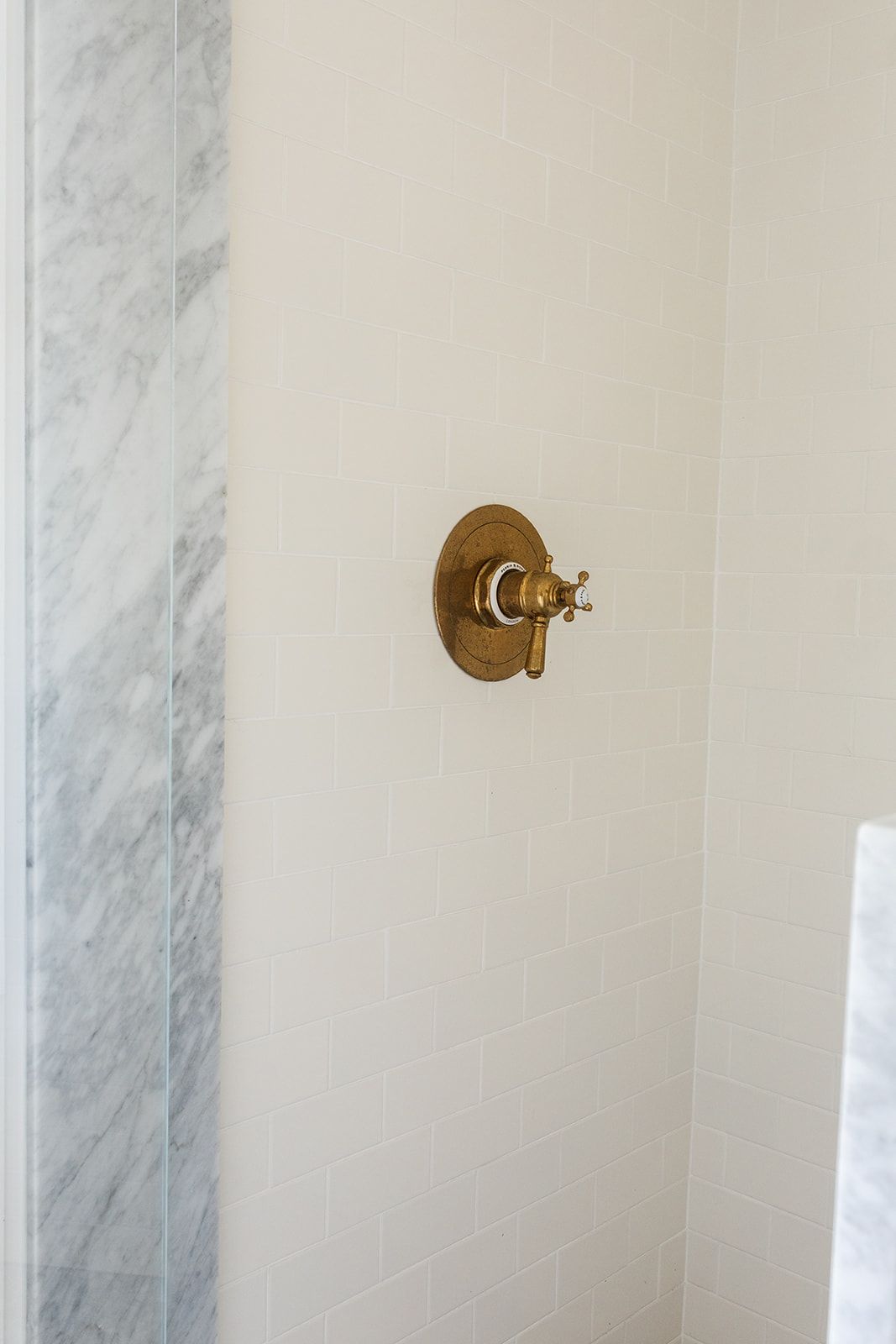 Antique brass cross-handle shower valve on white subway tile wall beside grey marble panel.