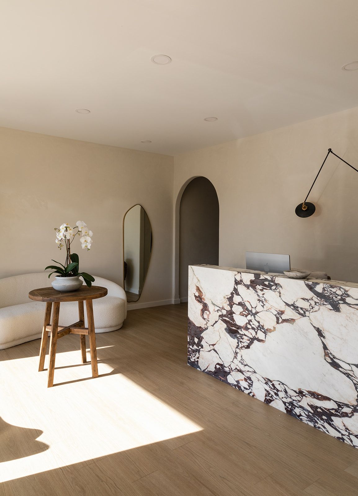 Modern interior space with marble reception desk, arched doorway, wooden stool with orchid, and architectural wall light.