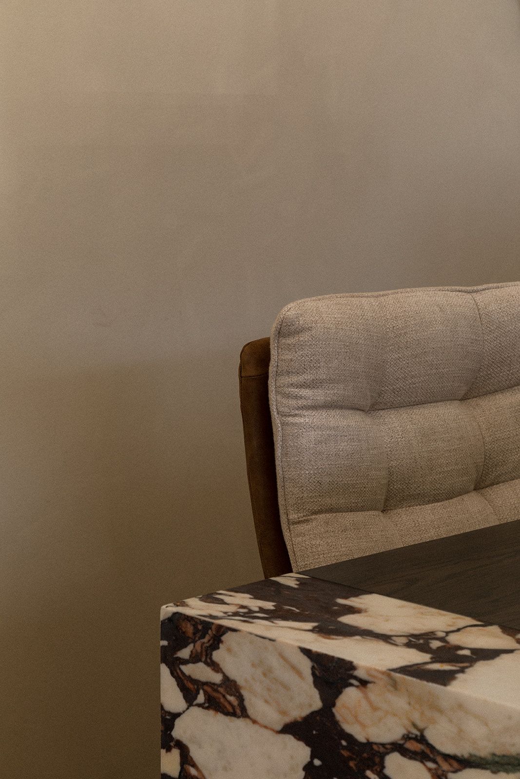 Modern gray upholstered chair with tufted back cushion sits against beige wall with marble-patterned detail below.