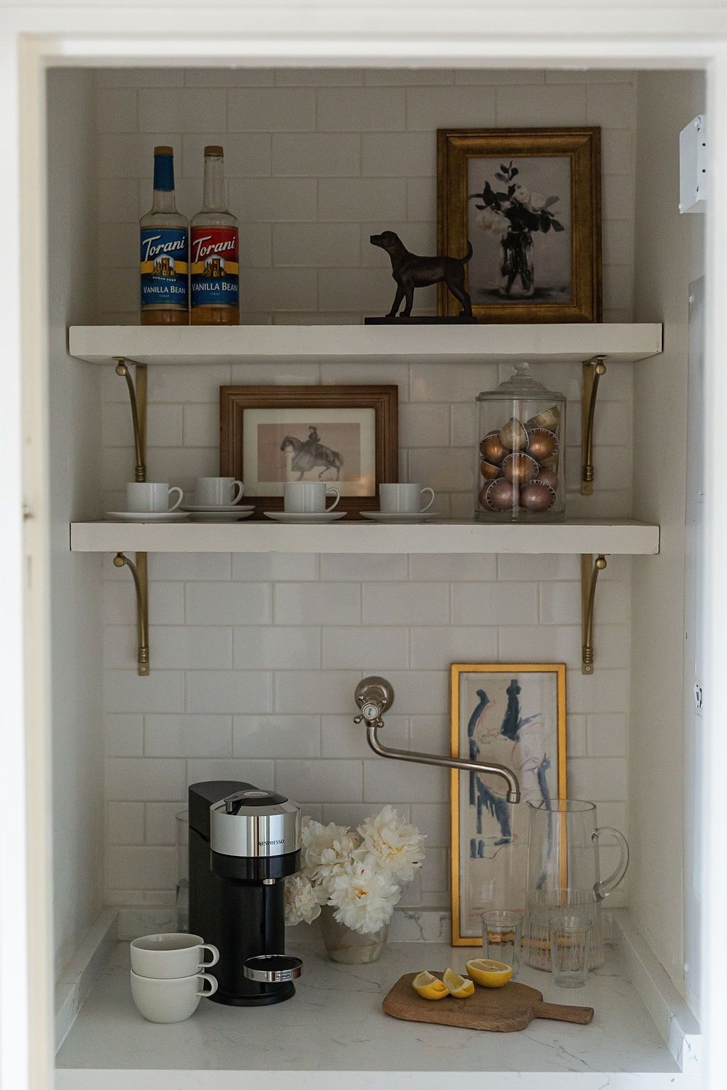 Stylish coffee station with white shelves featuring artwork, coffee maker, mugs, and decorative accessories on subway tile wall.