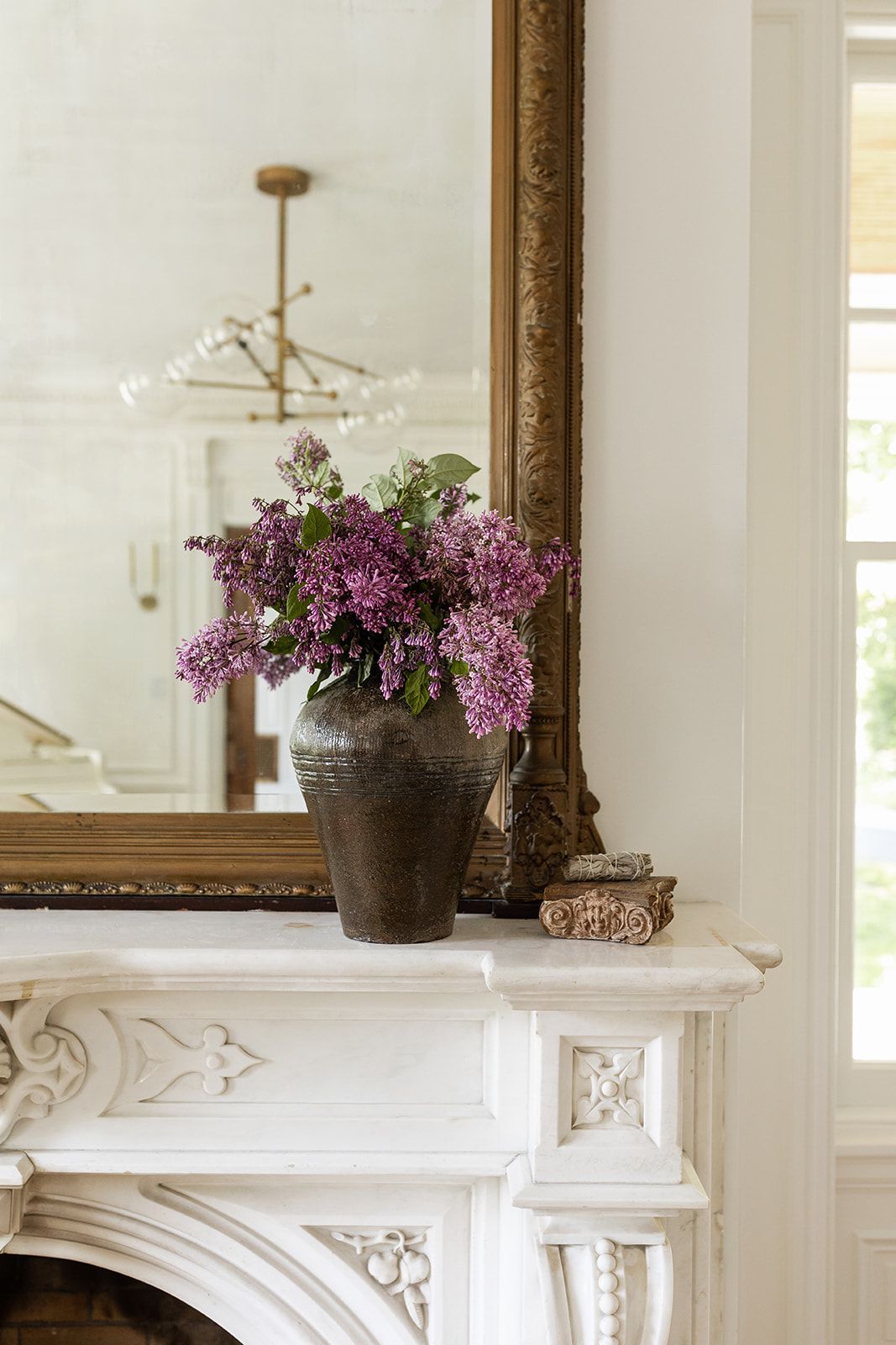 Purple lilac flowers in a dark vase on white marble mantelpiece with ornate gold mirror.
