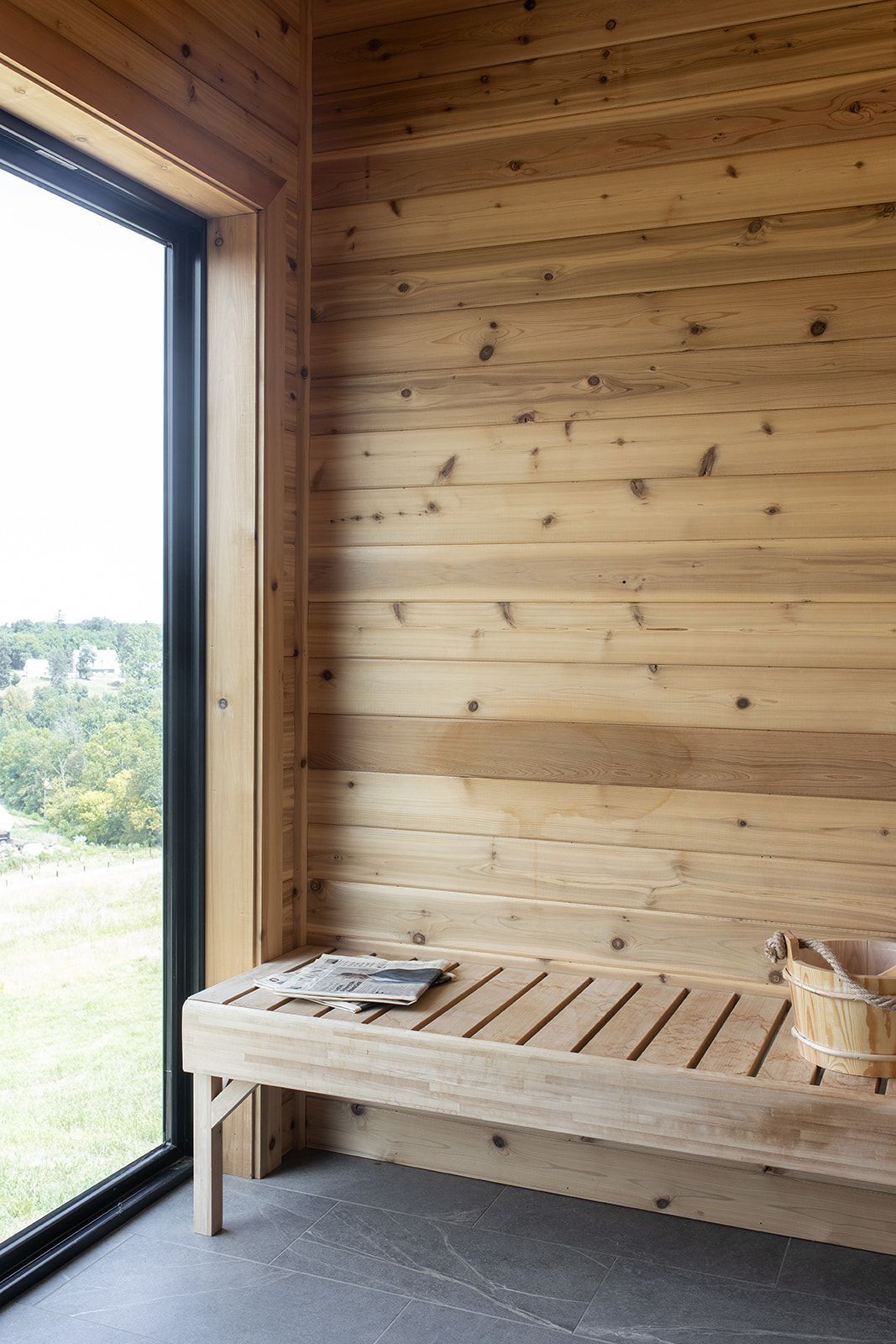 Modern sauna interior with cedar wood walls, slatted bench, and large window overlooking a green landscape.