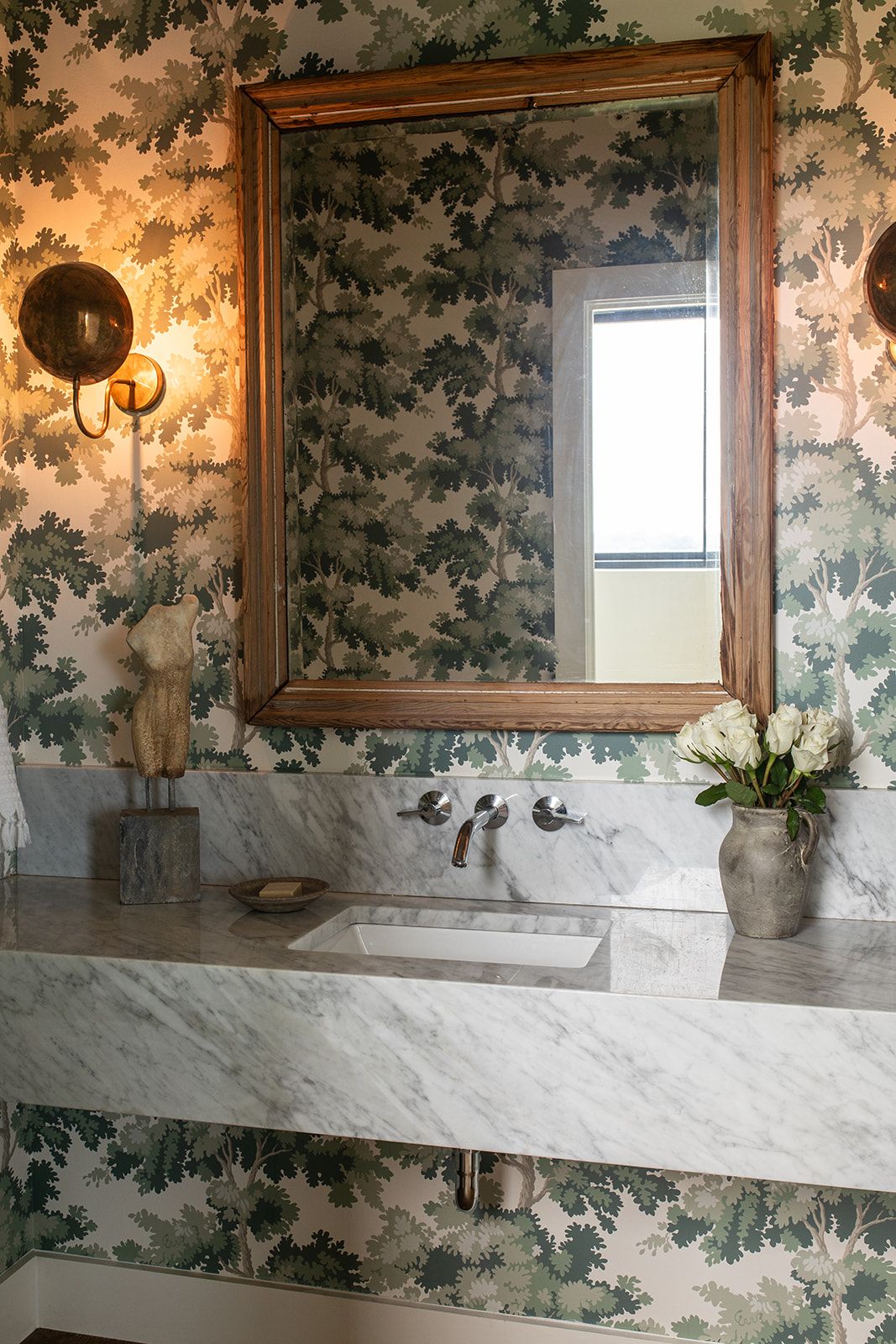 Elegant powder room with botanical wallpaper, marble vanity, wood-framed mirror, and brass wall sconces.