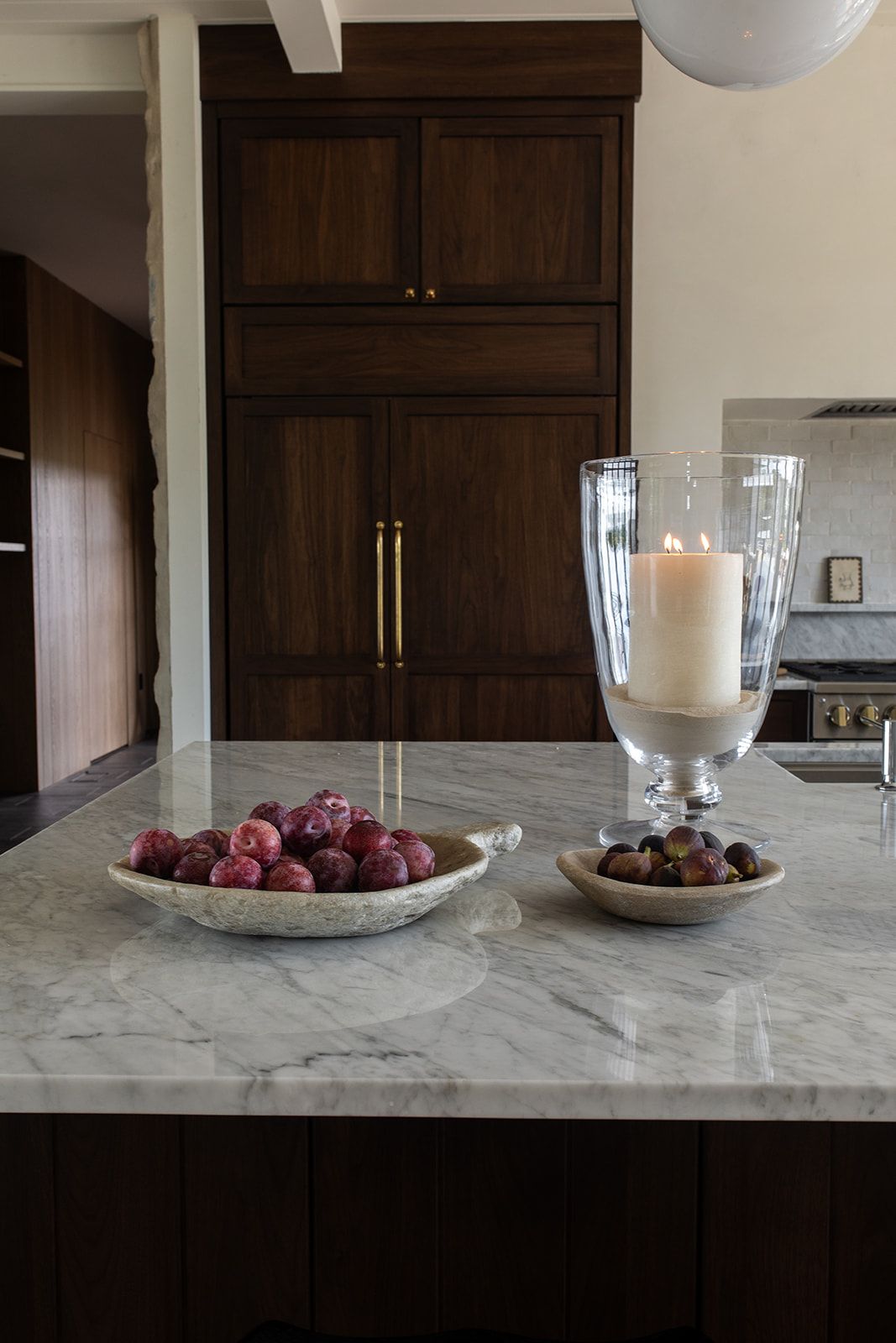 Marble kitchen island with fruit bowls and glass candle holder, dark walnut cabinetry with brass hardware in background.