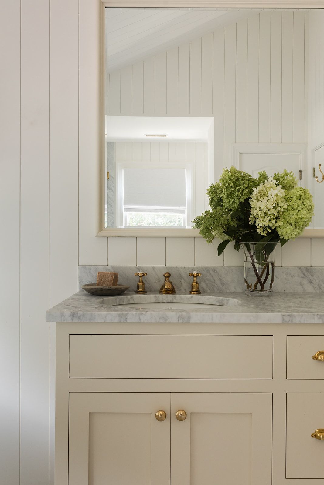 Elegant bathroom vanity with marble countertop, brass fixtures, white cabinetry, and fresh hydrangeas in a glass vase.