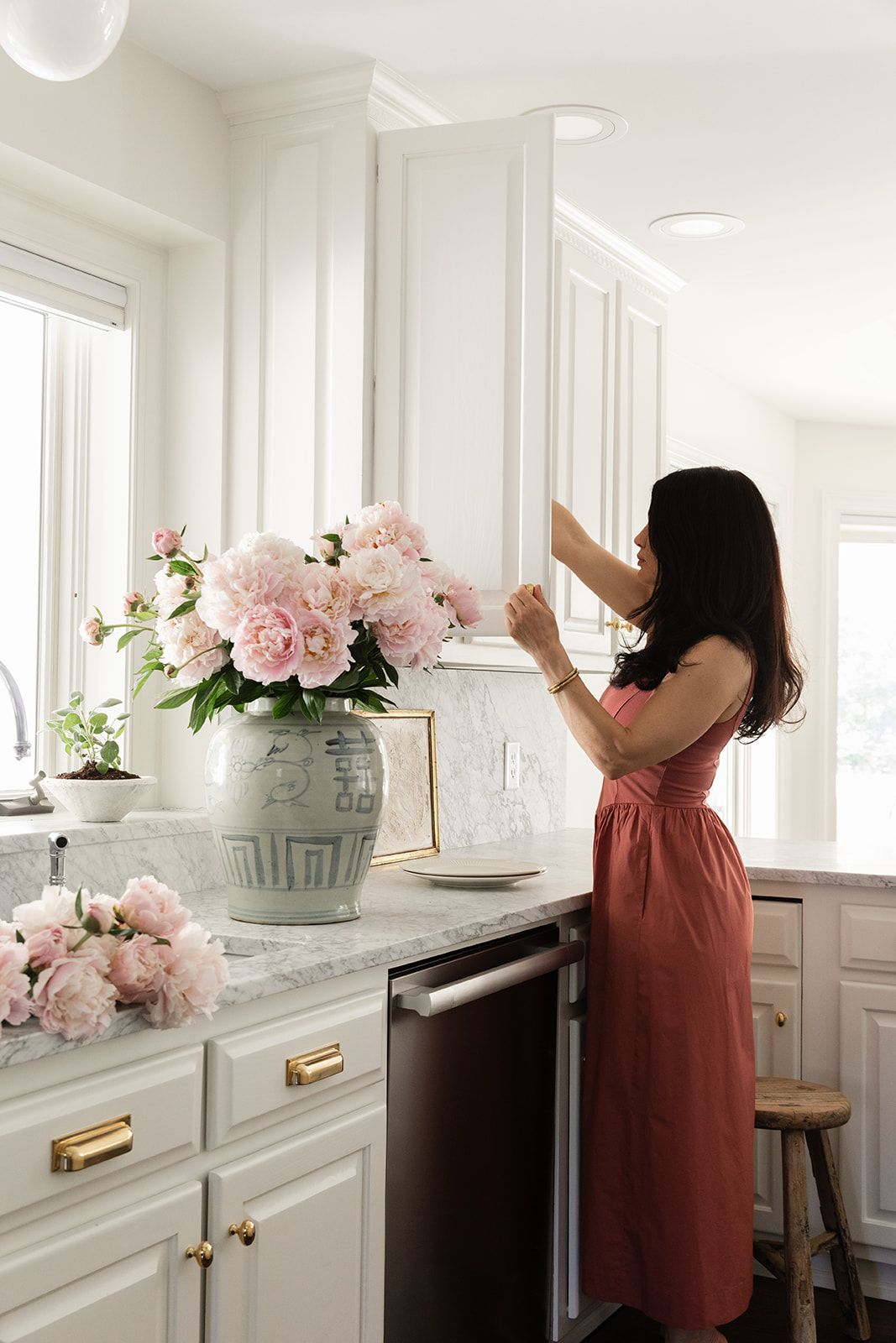 Woman in red dress arranging pink peonies in a green ginger jar vase in a bright white kitchen.