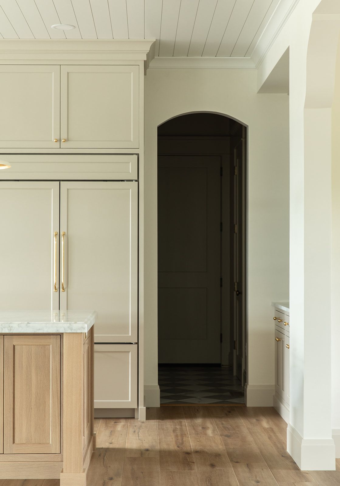 Neutral kitchen featuring greige cabinetry, brass pulls, wood floors, marble island, and an arched dark pantry doorway.