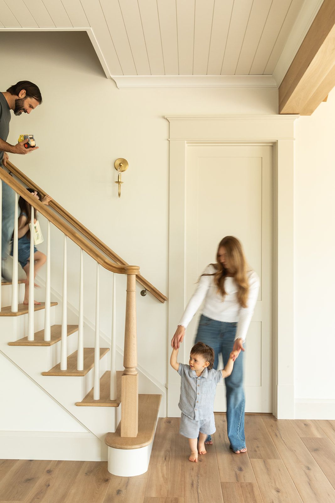 Family in a bright home entryway with wood staircase, mother helping toddler walk on hardwood floors.