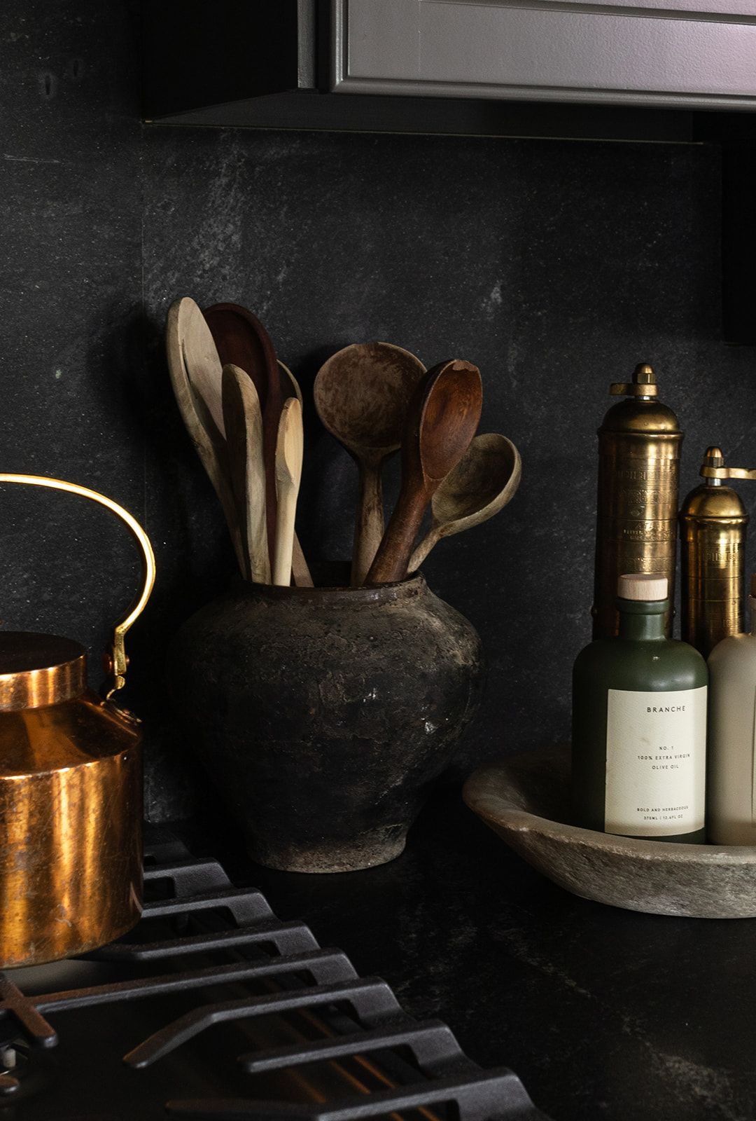 Vintage copper kettle, wooden utensils in clay pot, and glass bottles arranged on dark rustic kitchen shelf.