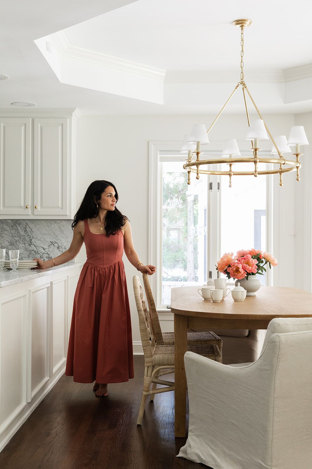 Woman in red dress standing in elegant white kitchen with gold chandelier, marble countertops, and round dining table.
