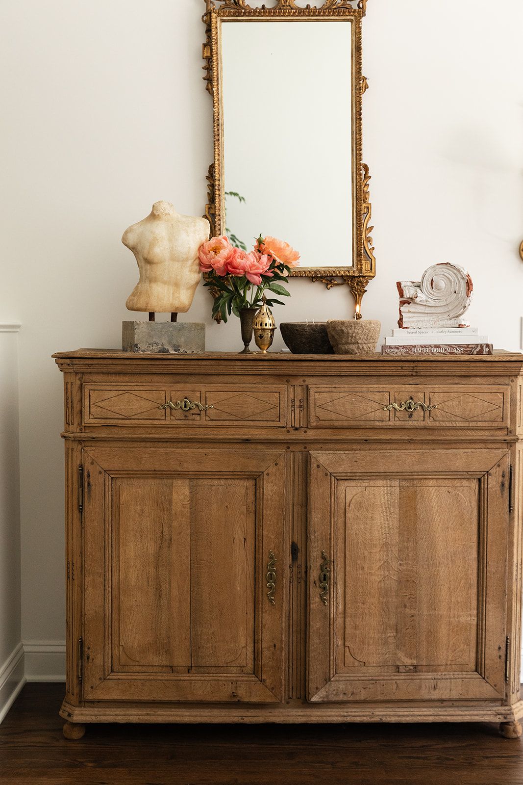 Antique wooden sideboard with gold mirror, pink flowers, and decorative objects in an elegant interior.