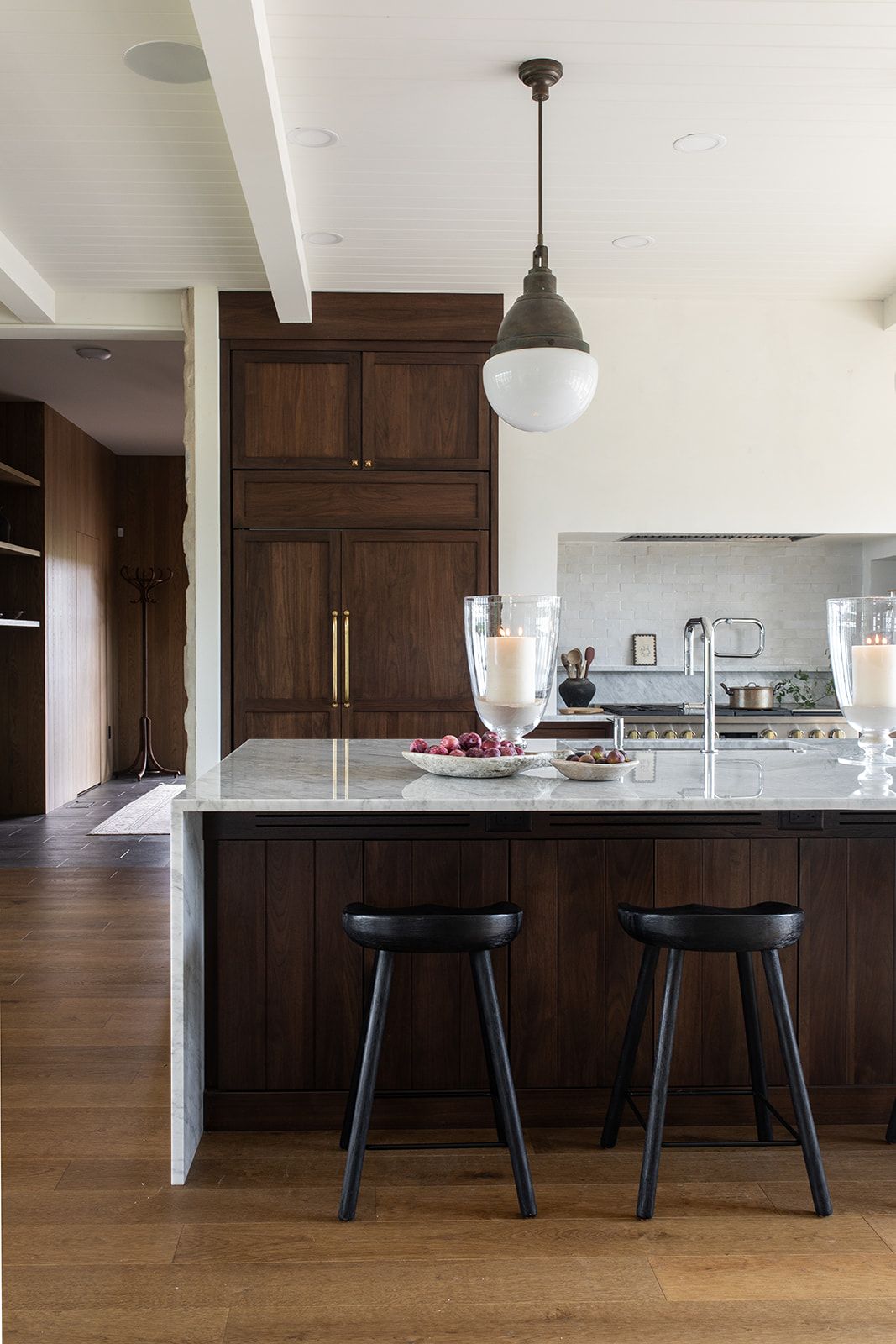 Dark walnut kitchen cabinets with marble island, black bar stools, and pendant light on hardwood floors.