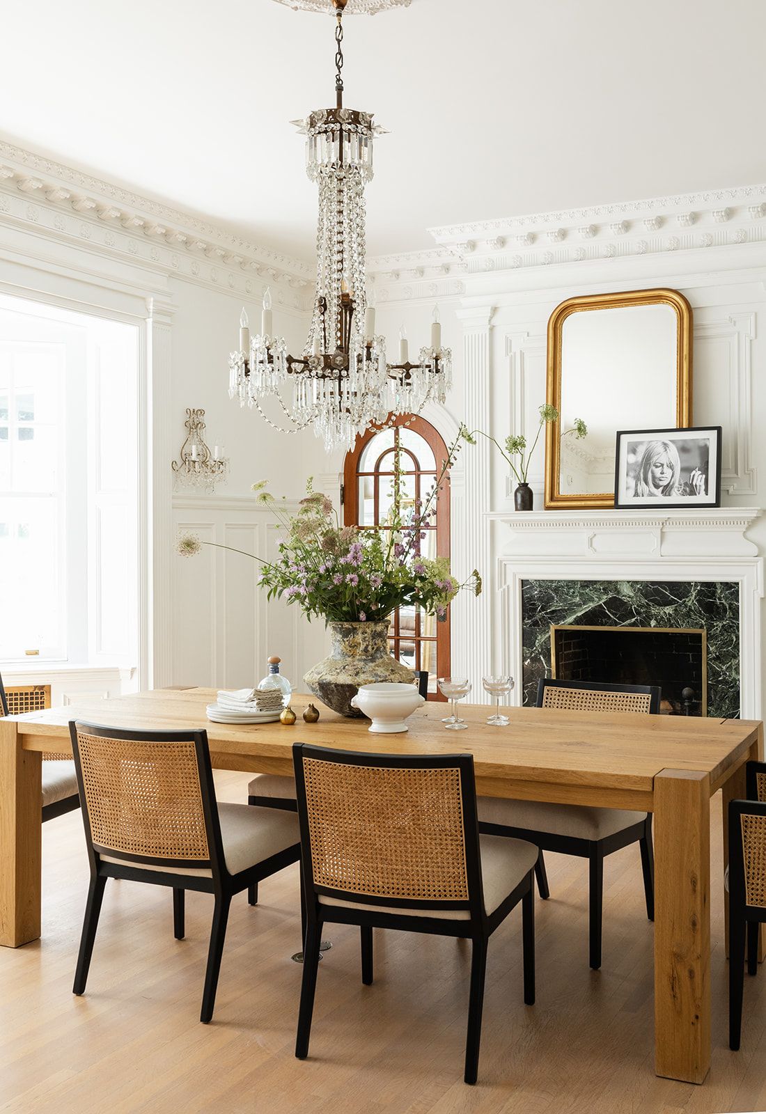 Elegant dining room with crystal chandelier, wooden table, cane chairs, and marble fireplace in bright white interior.