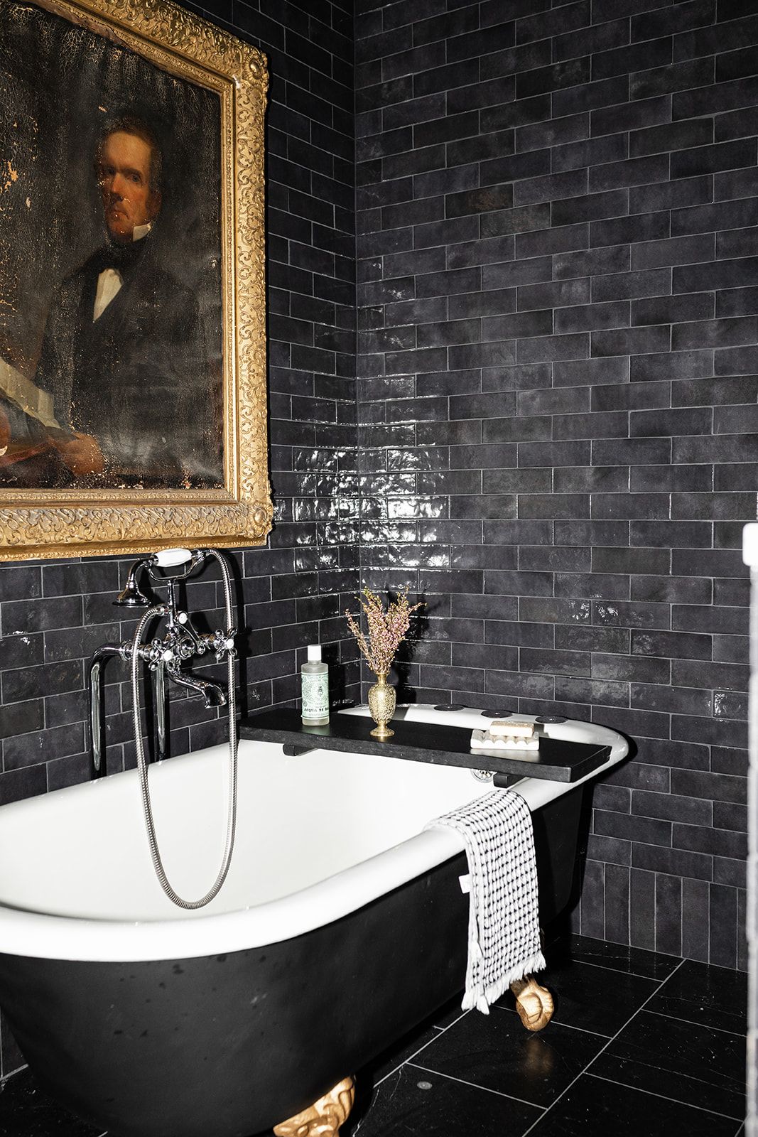 Dramatic black bathroom with vintage portrait, dark tile walls, and clawfoot bathtub.