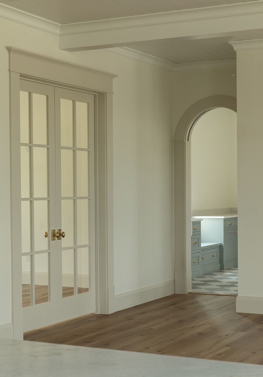 Elegant hallway with white French doors, brass hardware, arched doorway, and hardwood floors leading to a blue kitchen.