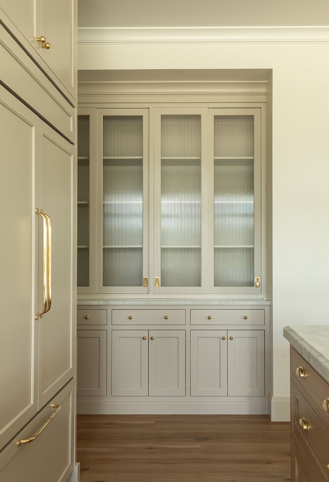 Built-in sage green china cabinet with reeded glass doors, brass pulls, and lower drawers in a classic kitchen pantry.