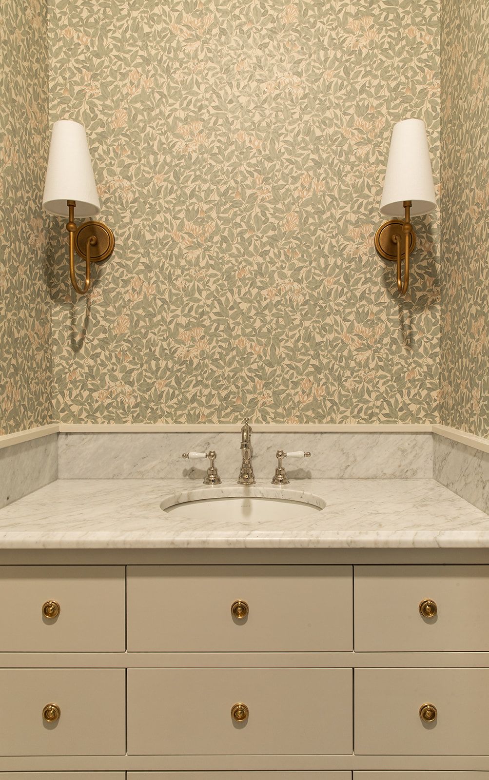 Elegant powder room with floral wallpaper, marble vanity, white cabinets with brass hardware, and gold wall sconces.