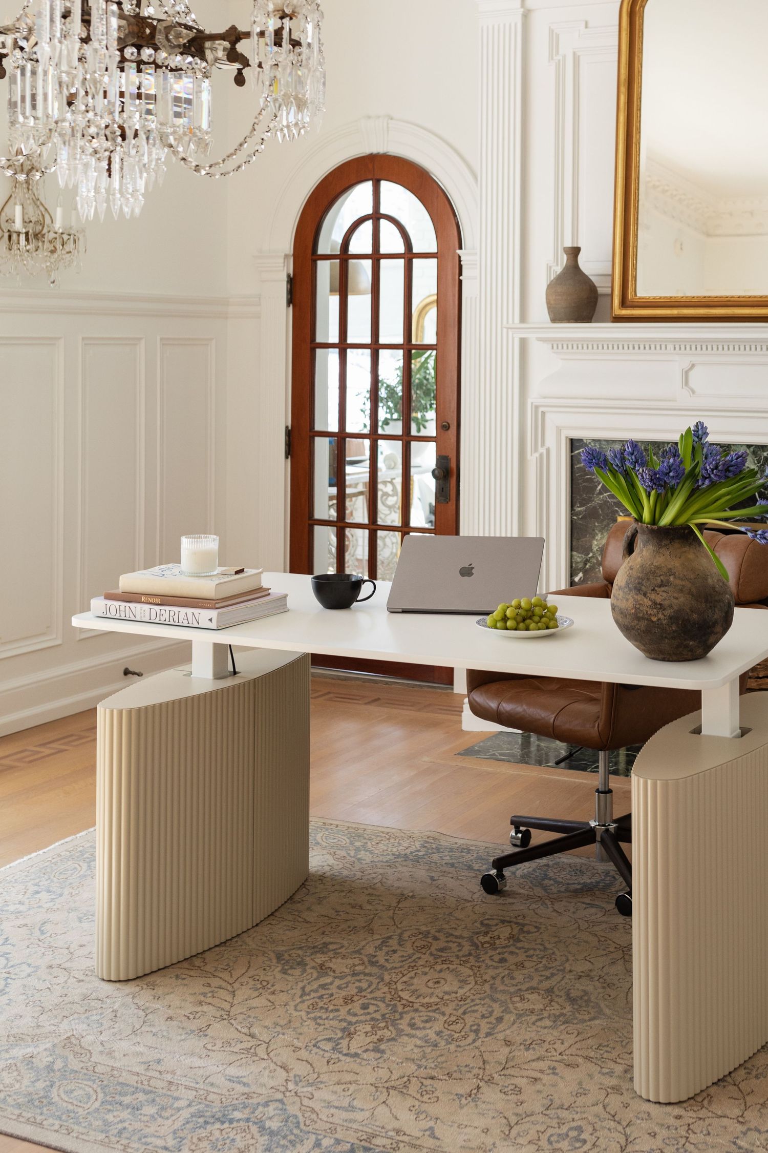 Elegant home office with a ribbed beige desk, leather chair, crystal chandelier, arched mirror, and blue hyacinths by a white fireplace.