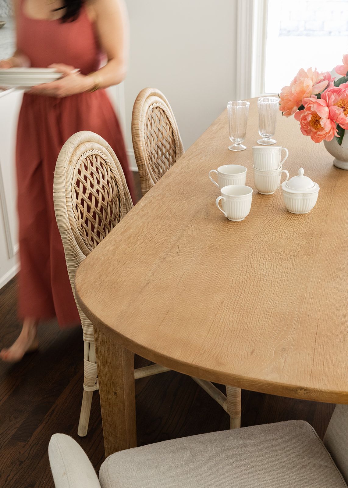 Oval wood dining table with rattan chairs, white tea set, and pink flowers, woman in red dress in background.
