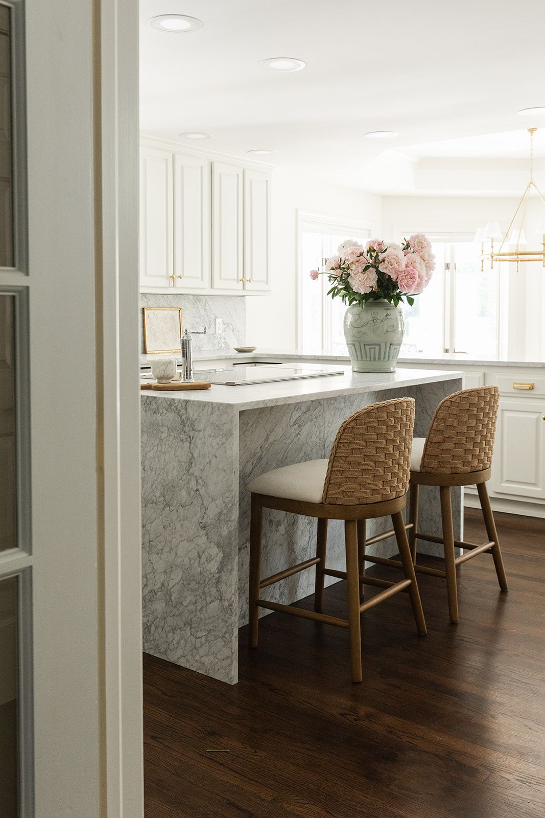 White kitchen with marble island, woven bar stools, pink peonies in vase, white cabinets, and dark hardwood floors.