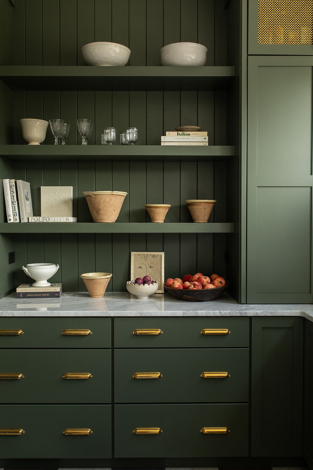 Green kitchen cabinetry with gold hardware, marble countertop, open shelves with bowls and glassware, and a bowl of red apples.