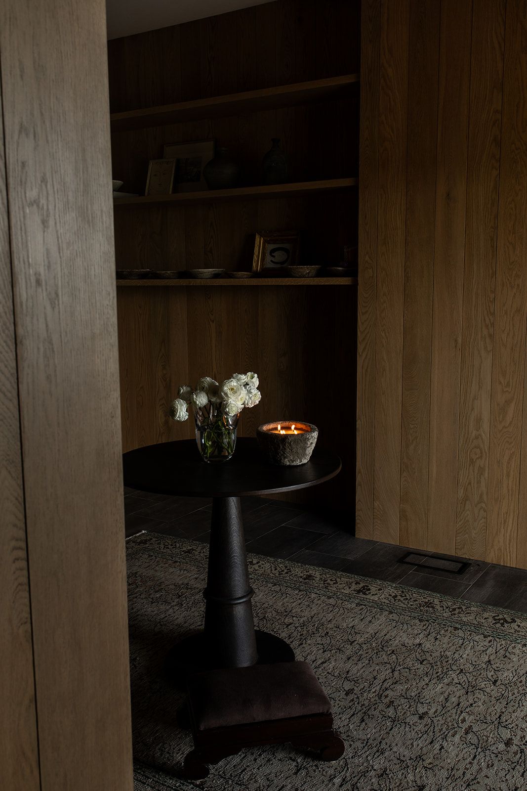 Dark moody interior with wooden shelves, a black pedestal table holding white flowers, and a glowing candle on a stone surface.