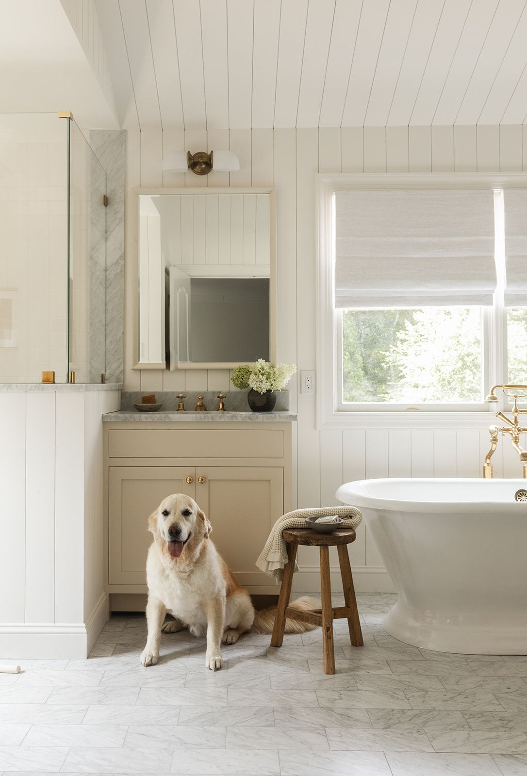 White farmhouse bathroom with golden retriever, freestanding tub, beige vanity, marble floors, and shiplap walls.
