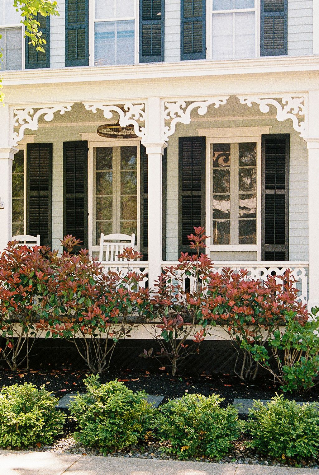 A white Victorian house with ornate trim features a welcoming front porch adorned with flowering bushes and landscaping.