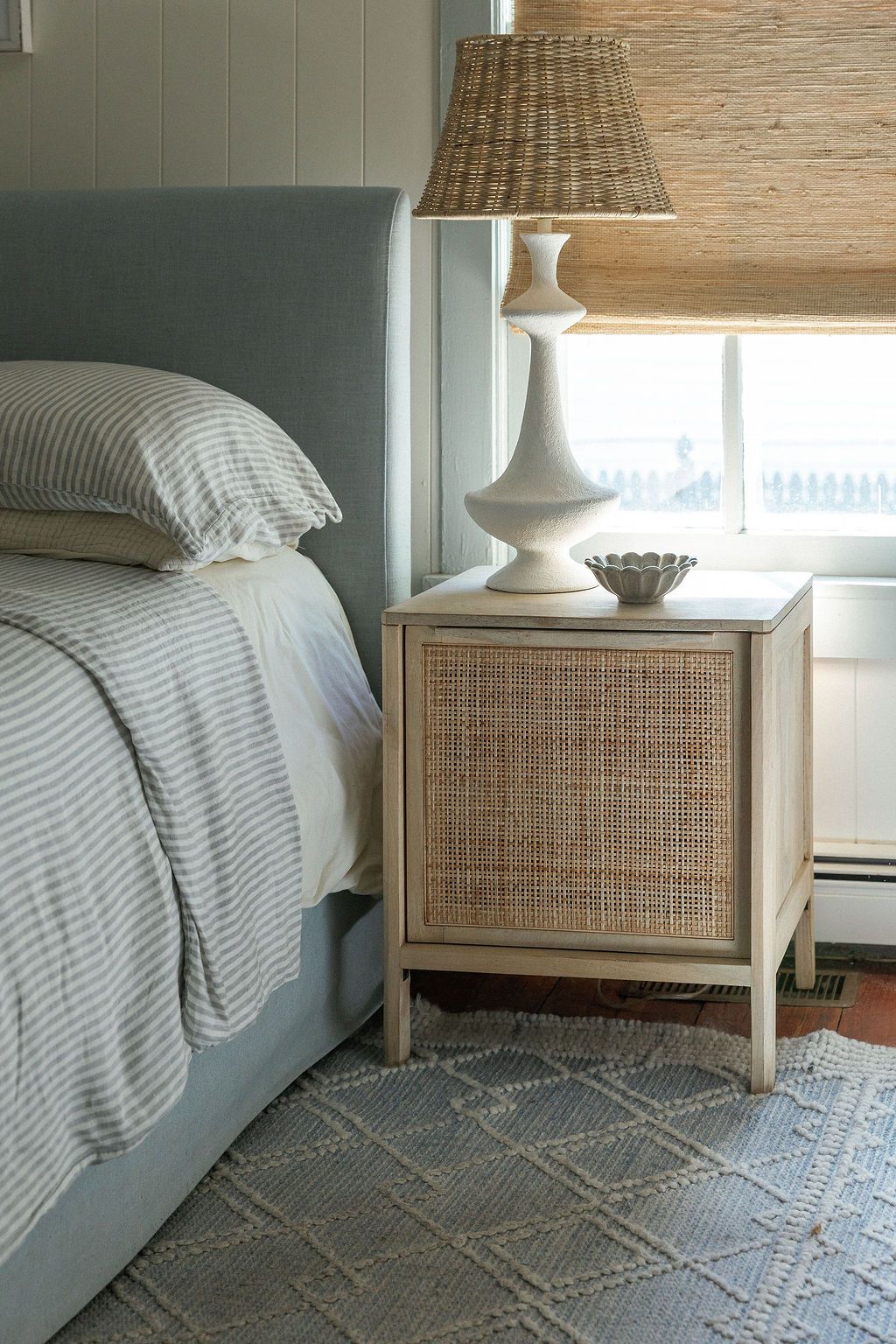 Modern gray bedroom with rattan nightstand, white table lamp, striped bedding and geometric patterned area rug.