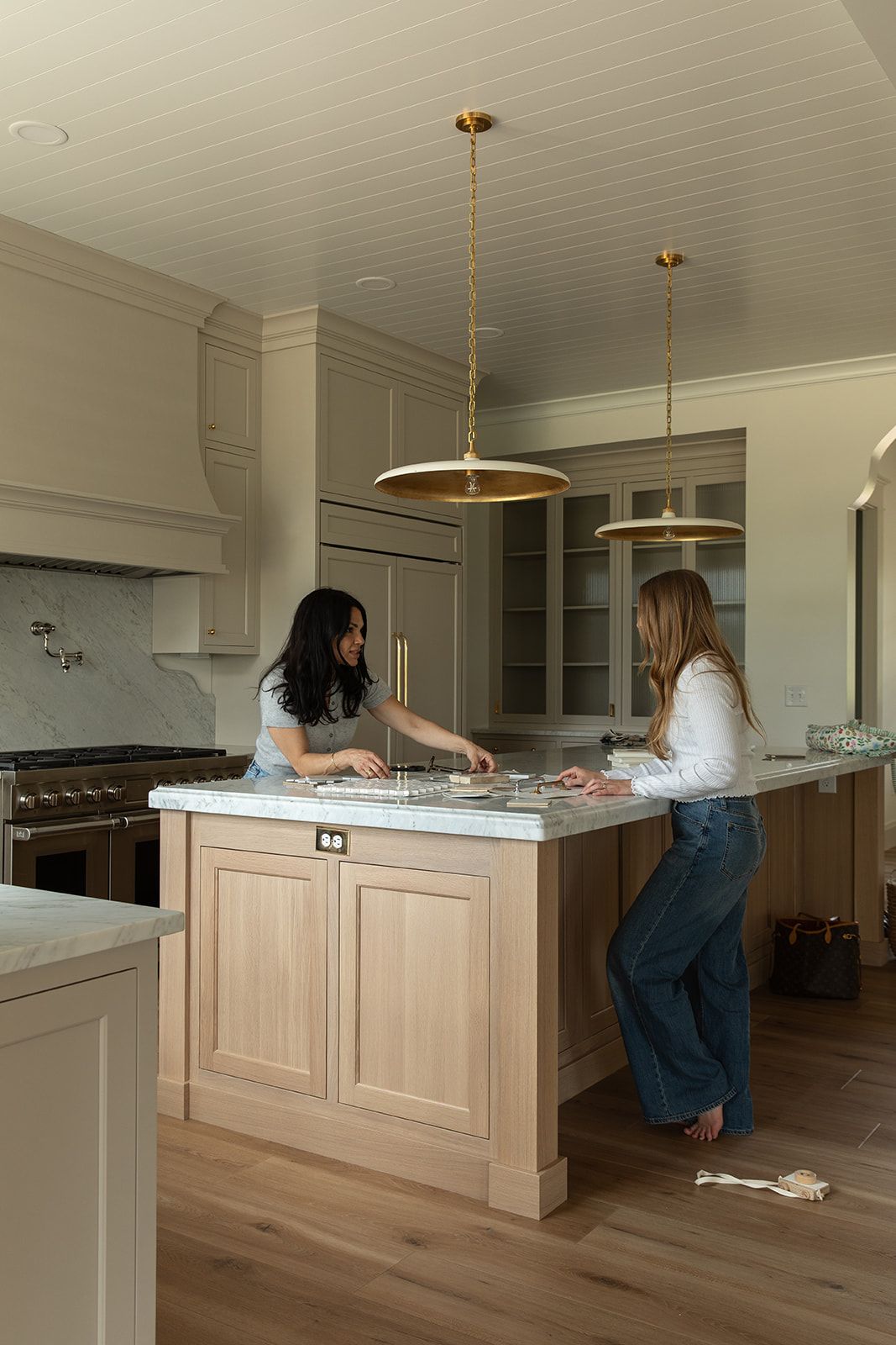 Two women working at a light wood kitchen island with marble countertop, brass pendant lights, and sage green cabinetry.
