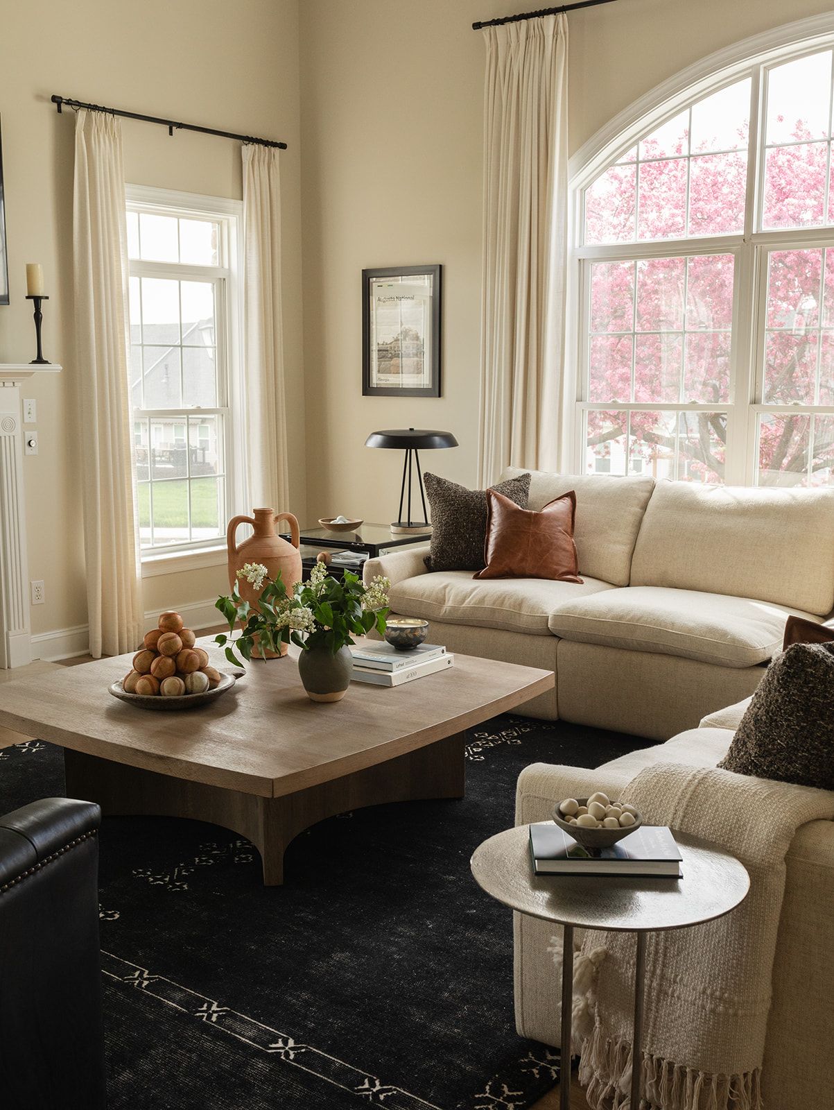 Cozy living room with cream sectional sofa, wooden coffee table, and large windows showcasing pink flowering trees outside.
