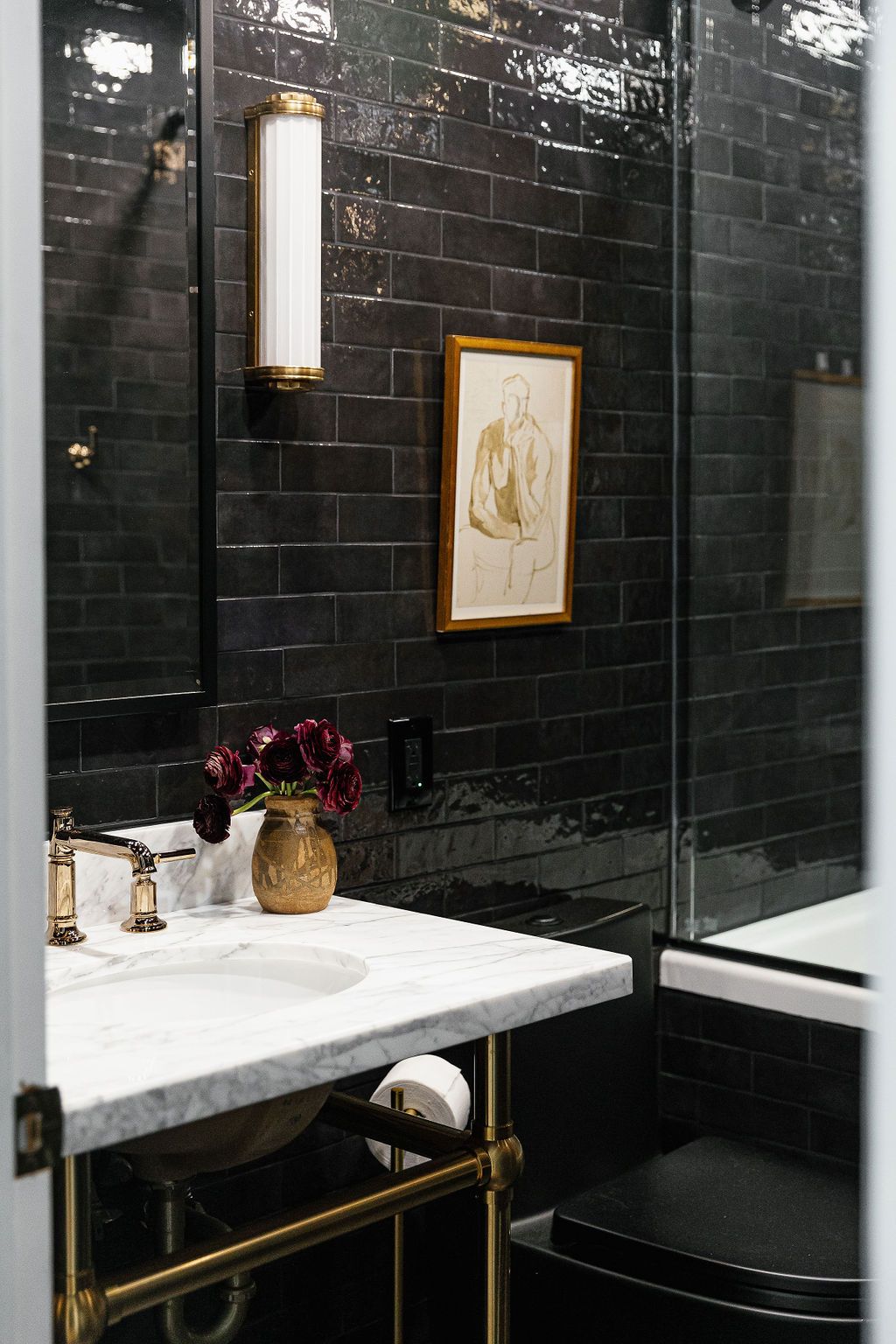 Elegant bathroom interior with black subway tile walls, white marble counters, brass fixtures and framed wall art.