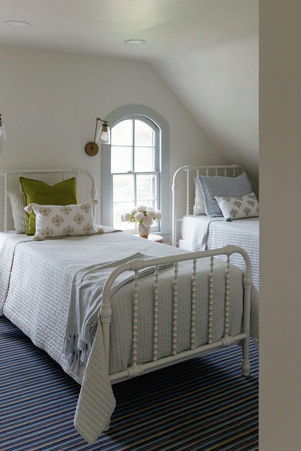 Cozy attic bedroom with white iron bed frame, striped rug, and arched window featuring green accent pillows.