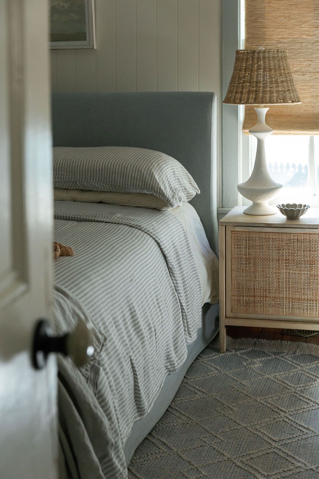 Cozy bedroom interior with upholstered bed, striped bedding, woven table lamp and rattan nightstand in neutral tones.