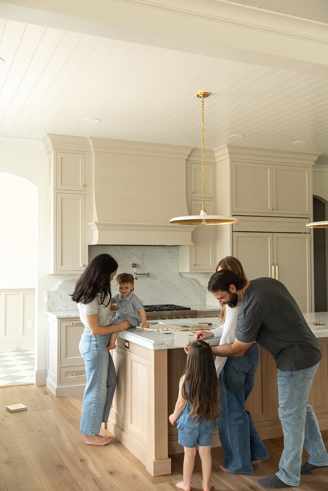 Family of four gathered around a kitchen island in a bright, modern kitchen with cream cabinets and gold accents.