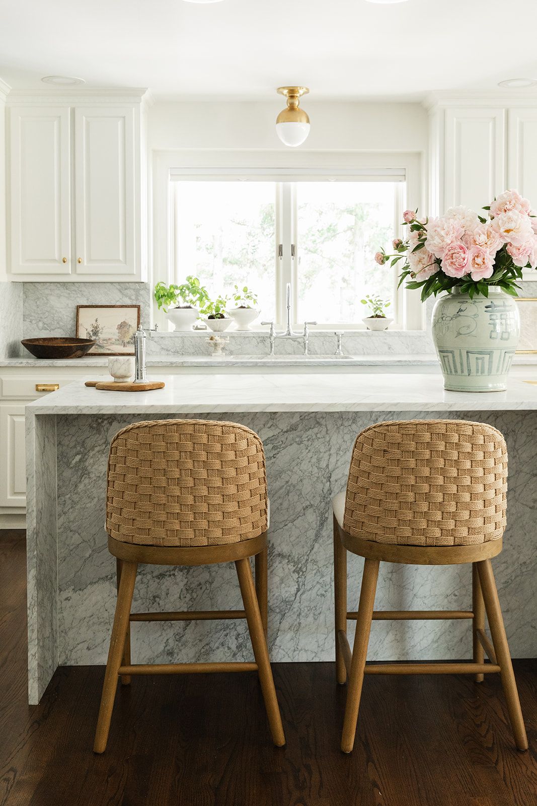 White kitchen with marble island, woven bar stools, white cabinets, brass fixtures, and pink peonies in a vase.
